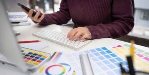 Was kostet ein Logo Design? Frau sitzt am Computer. Man sieht die Hände auf der Tastatur. Drum herum liegen Farbbücher mit Farbpaletten