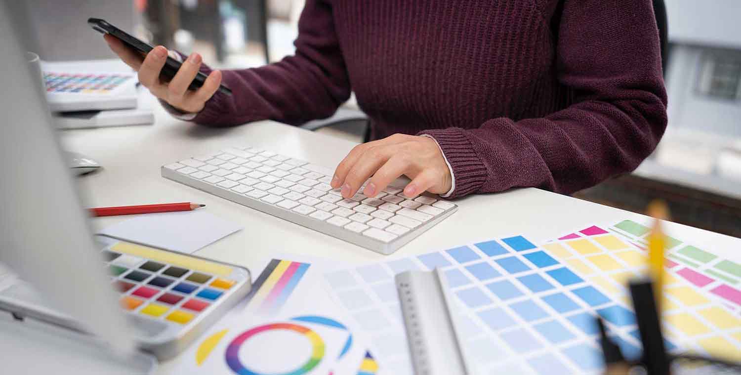Was kostet ein Logo Design? Frau sitzt am Computer. Man sieht die Hände auf der Tastatur. Drum herum liegen Farbbücher mit Farbpaletten